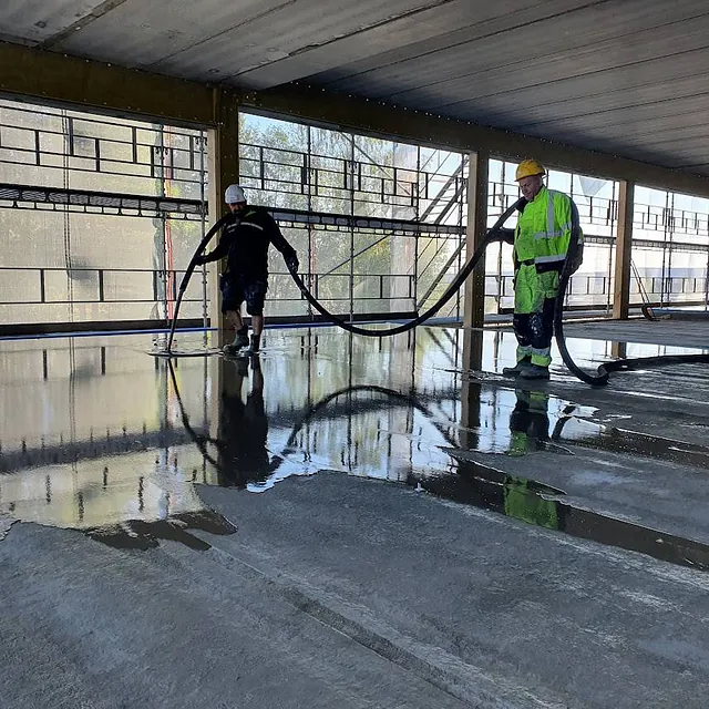 To arbeidere fordeler flytende avrettingsmasse i et åpent bygg med store vinduer